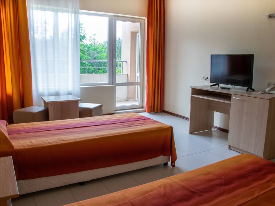 Habitació d'hotel amb dos llits, una taula de nit amb televisió, i un balcó amb cortines taronges.