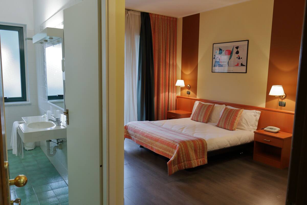 Habitación de hotel con cama, mesitas de noche, lámpara, obra de arte en la pared y baño con lavamanos y espejo.