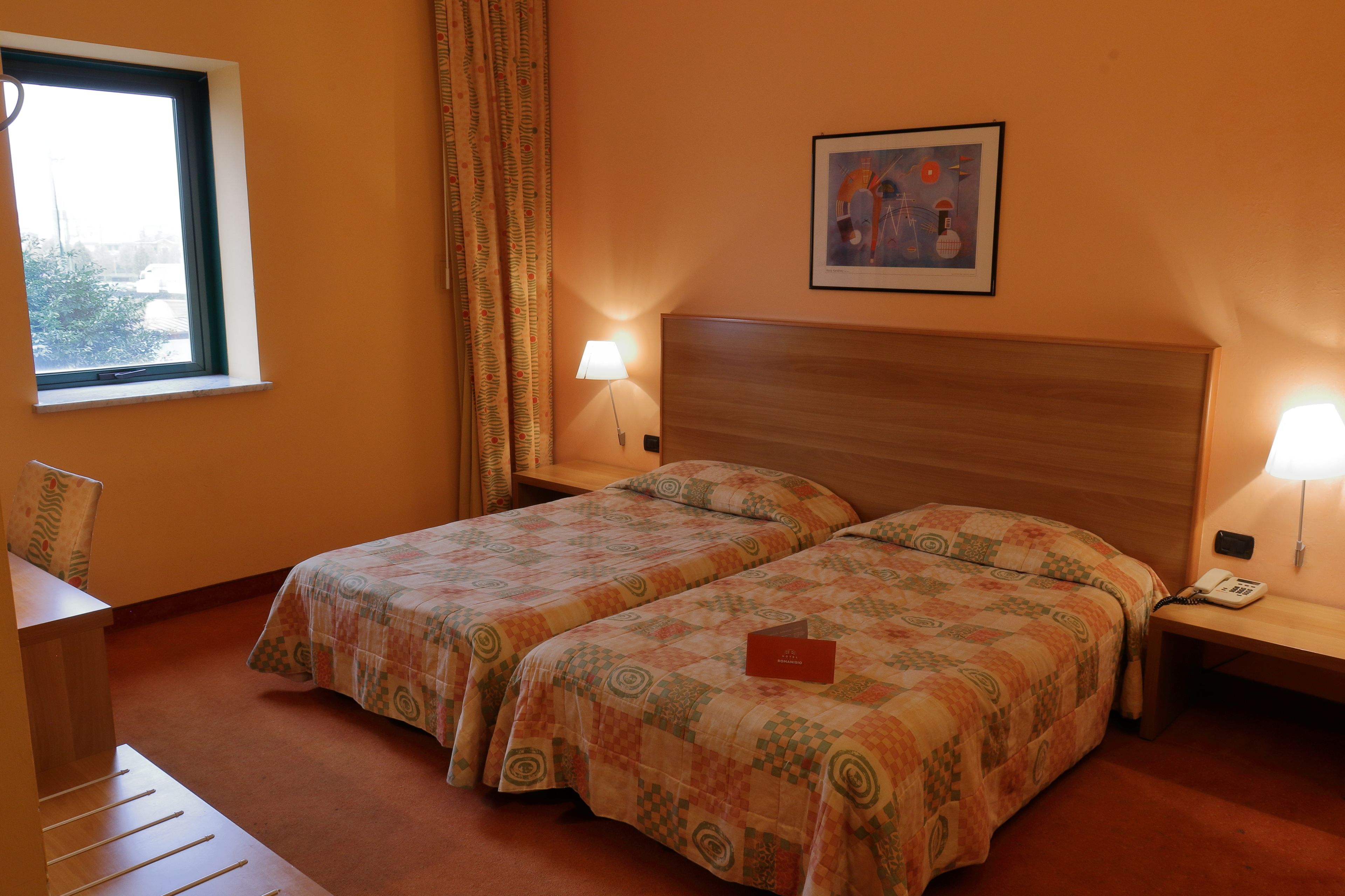 Ein Hotelzimmer mit zwei Betten, einem Schreibtisch, einem Stuhl, einem Fenster, einem Gemälde und einem Telefon auf dem Nachttisch.