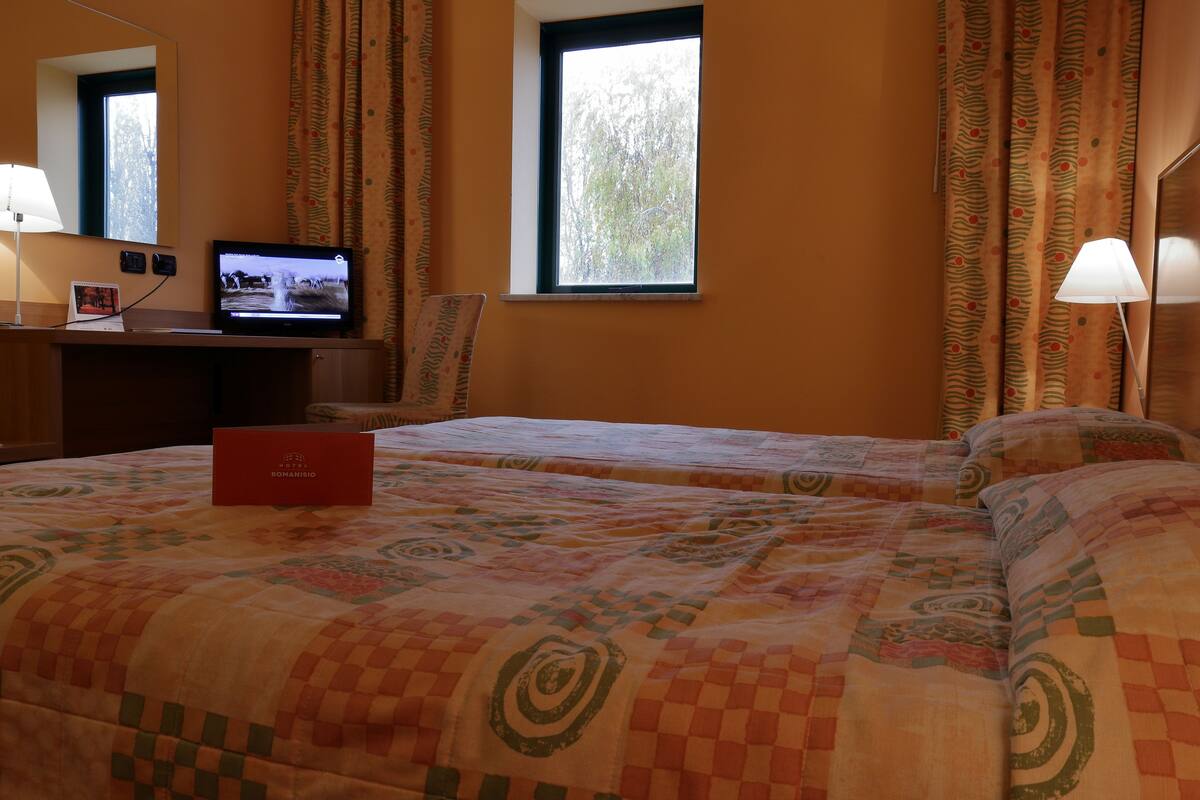 Habitación de hotel con dos camas, televisión, escritorio con computadora y ventana con cortinas.