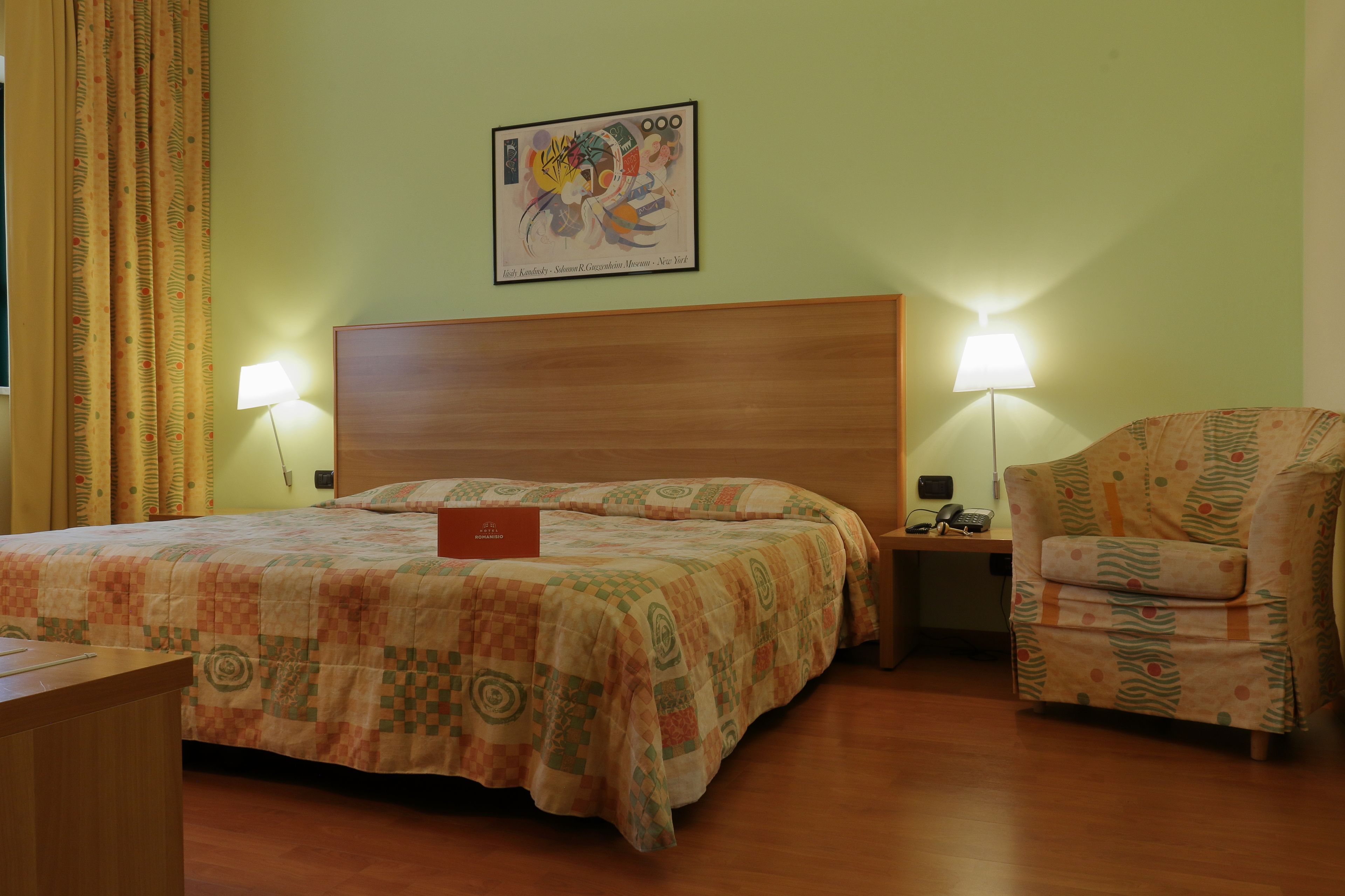 A hotel room with a large bed, a wooden headboard, two bedside lamps, a patterned armchair, and a framed artwork on the wall.