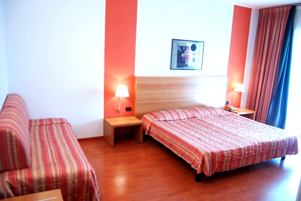 Habitación de hotel con cama, dos mesitas de noche, un sofá y un cuadro colgado en la pared.