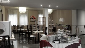 A dining area with tables set for a meal, chairs, a buffet table with food and drinks, and a kitchen area in the background. - Hotel Romanisio (Fossano)