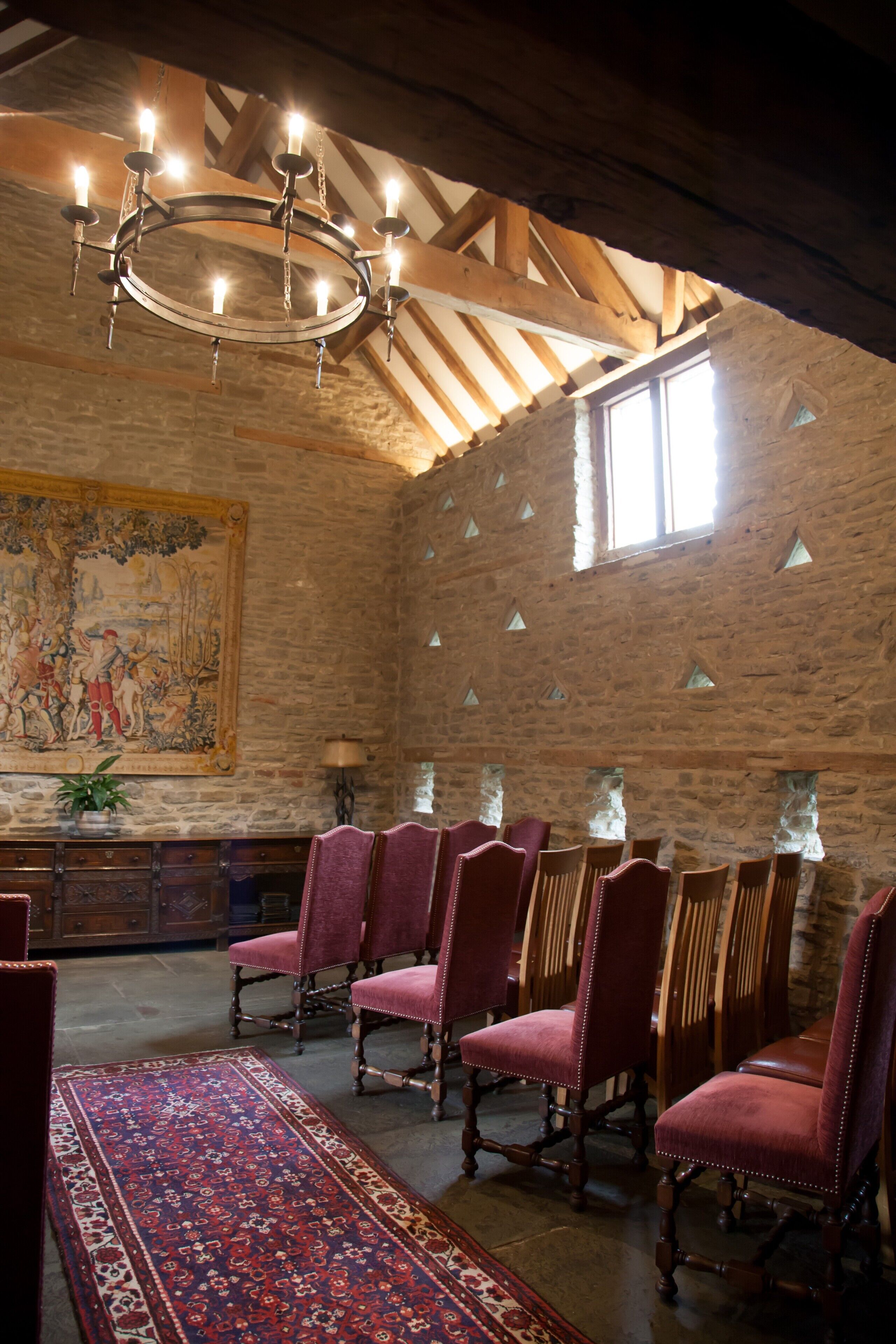 A room with a large wooden ceiling, stone walls, and a chandelier. There are several chairs and a table with a rug.