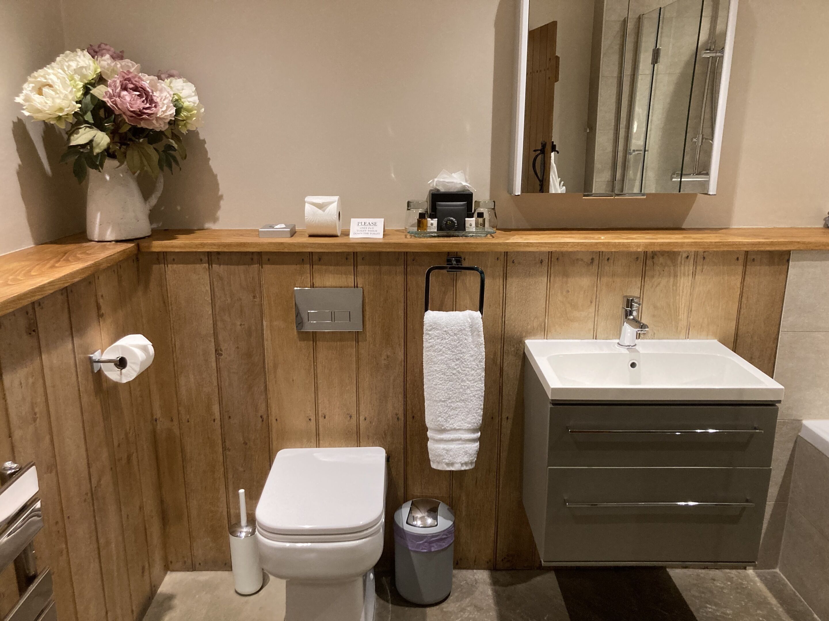 Une salle de bain avec un mur recouvert de panneaux en bois, des toilettes blanches, un lavabo avec un miroir et un vase de fleurs posé sur le comptoir.