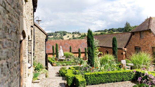 A courtyard with stone walls, a brick building, and a garden with various plants and a seating area. - Old Downton Lodge (Ludlow)
