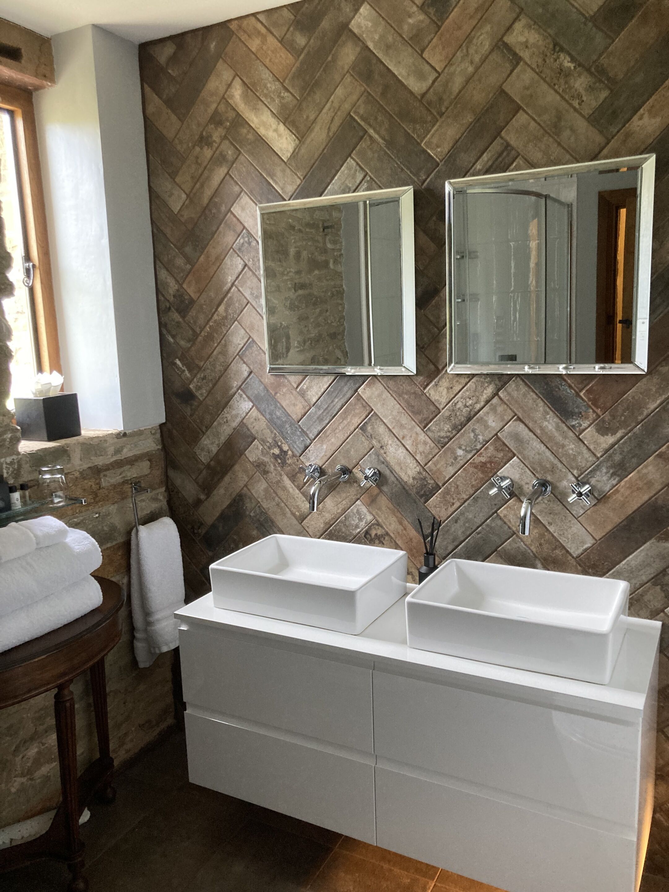 A bathroom with two white sinks, a wooden vanity, and a herringbone-patterned tiled wall.