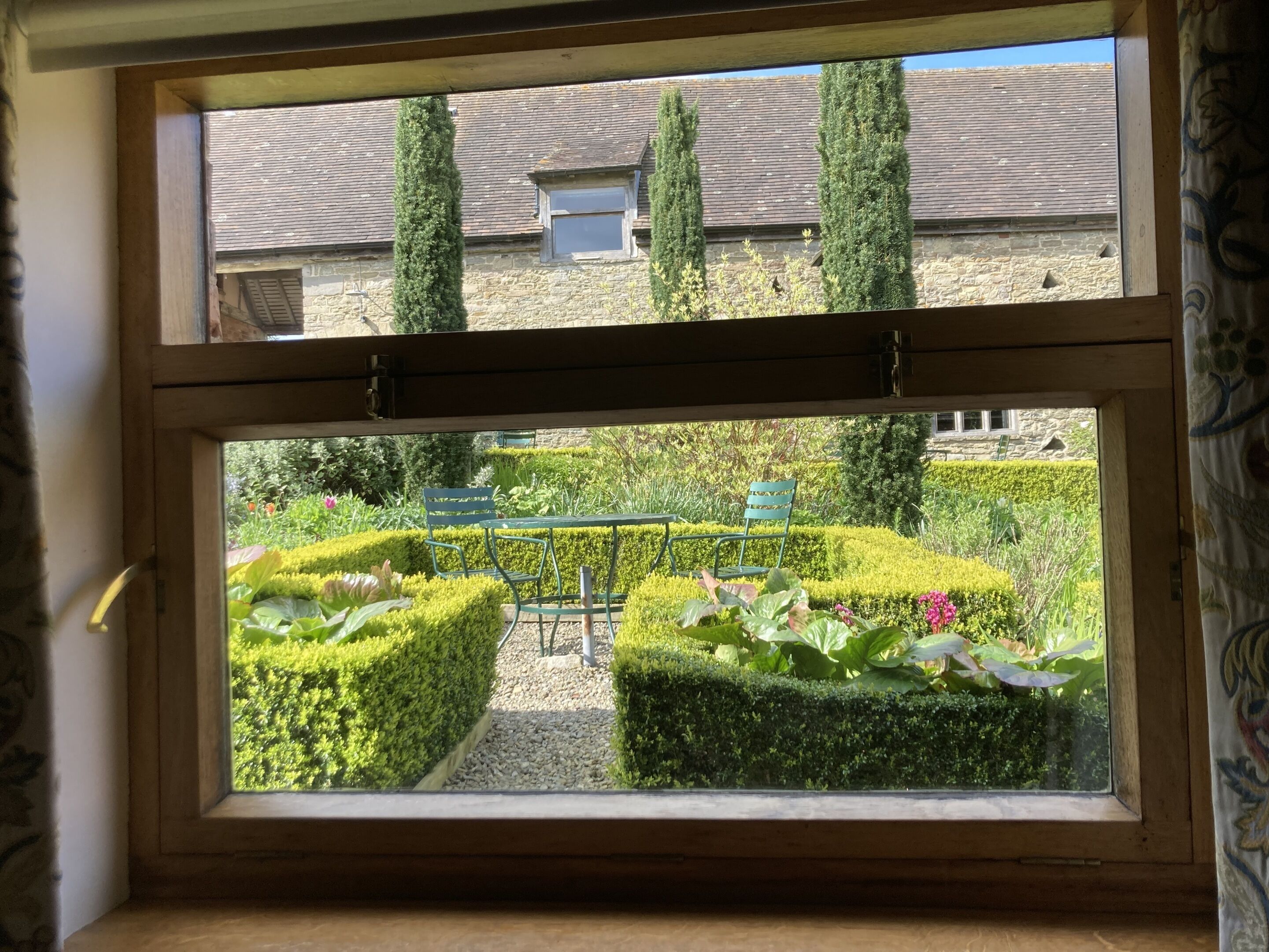 Una ventana con vista a un jardín cuidado, que incluye setos recortados y una mesa con sillas.