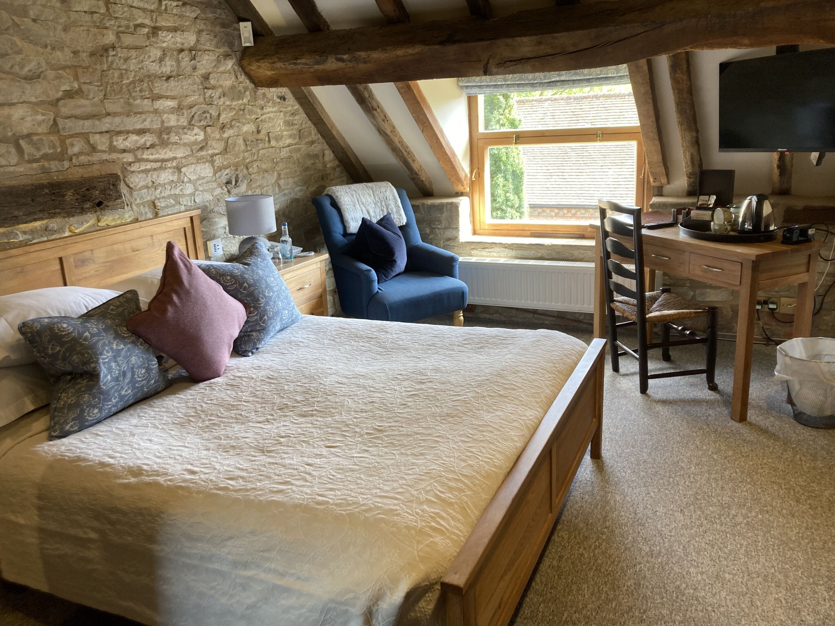 Ein Schlafzimmer mit Steinsturz, Holzbalkendecke, einem Bett mit gemusterten Kissen, einem blauen Sessel, einem Schreibtisch mit Stuhl und einem Fernseher.
