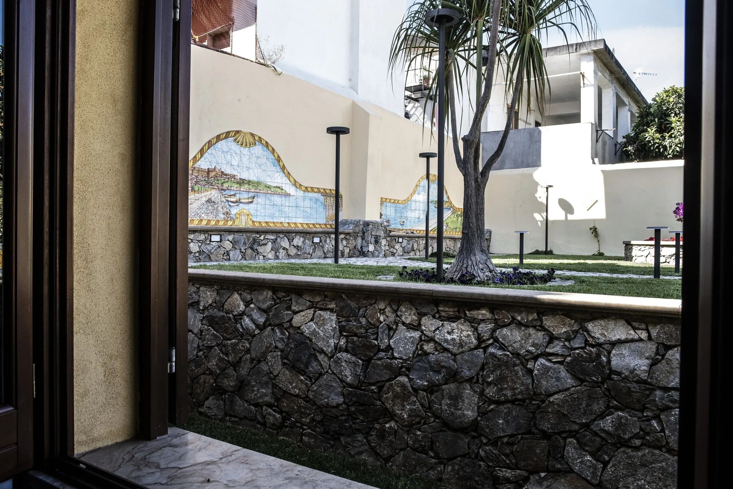 A stone wall with a mosaic mural, a palm tree, and a modern building in the background.