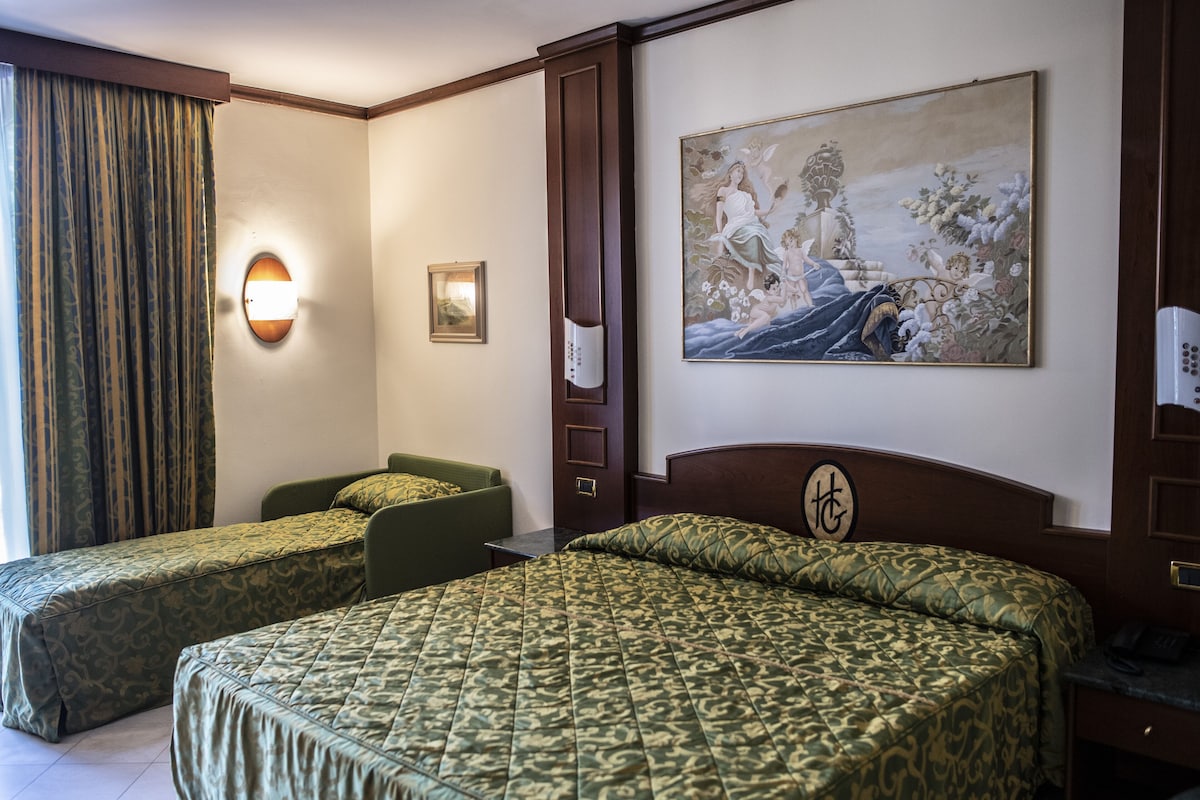 Una habitación de hotel con dos camas, un cuadro en la pared, una mesita de noche y una ventana con cortinas.