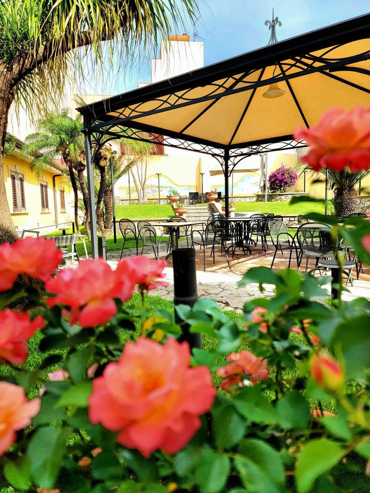 Eine Außenpatio-Terrasse mit Tischen und Stühlen, umgeben von Grünpflanzen und einem Gebäude im Hintergrund.