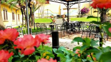 Eine Außenpatio-Terrasse mit Tischen und Stühlen, umgeben von Grünpflanzen und einem Gebäude im Hintergrund.