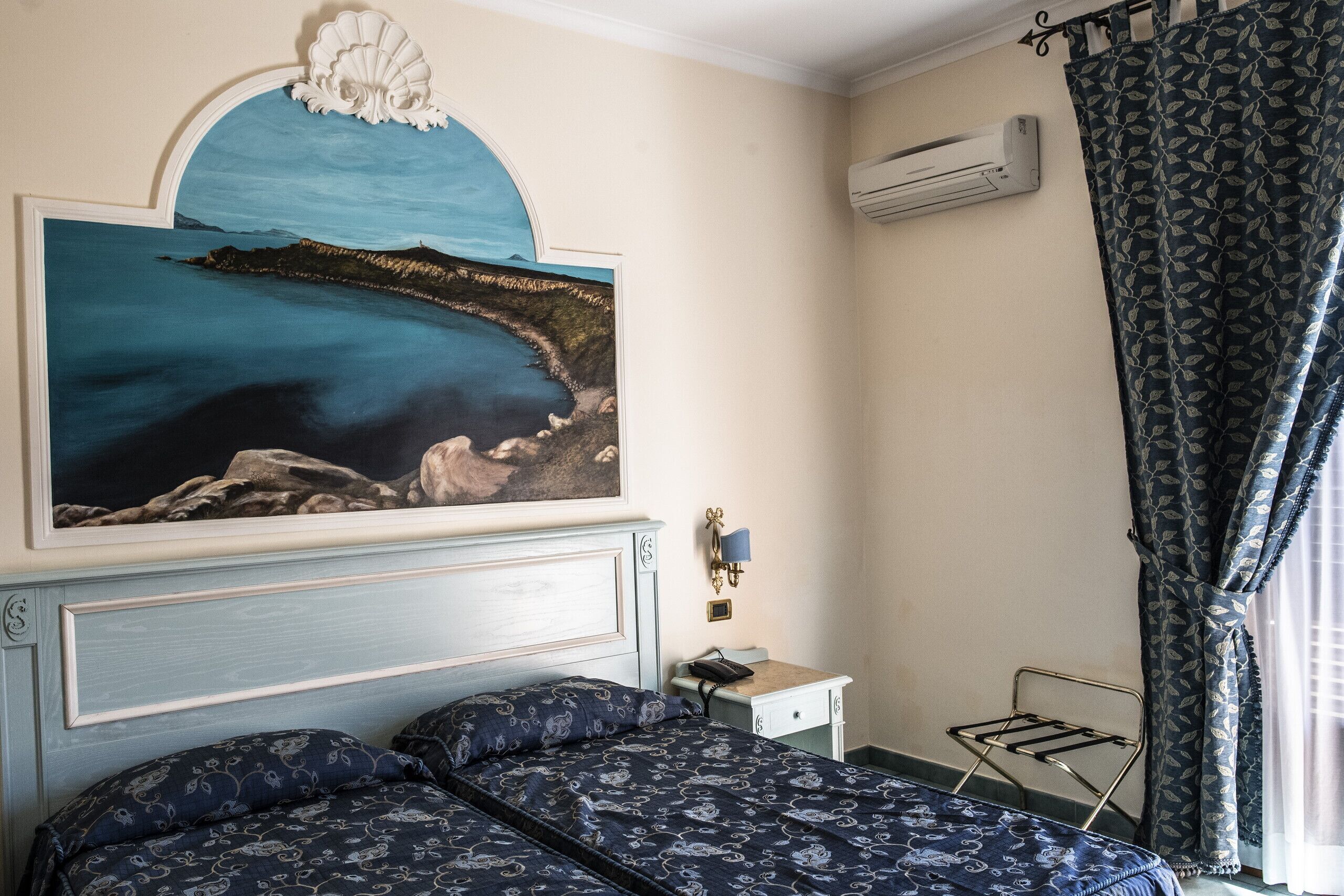 A bedroom with a bed, a nightstand, a wall-mounted lamp, an air conditioning unit, and a window with curtains.