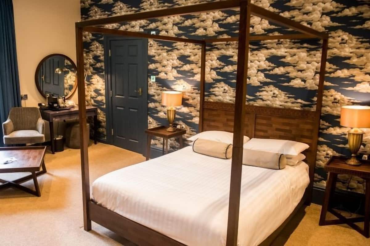Uma cama de dossel com estrutura de madeira, duas mesas de cabeceira com luminárias, uma cadeira, uma escrivaninha e uma porta com papel de parede estampado com nuvens.