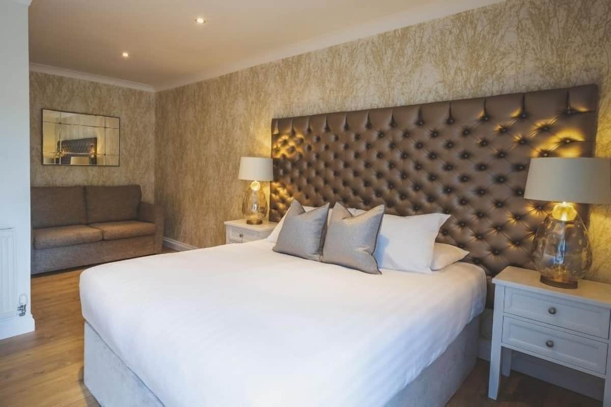 Quarto moderno com uma cama grande, cabeceira estofada, duas mesas de cabeceira com abajures, um sofá e um espelho na parede.