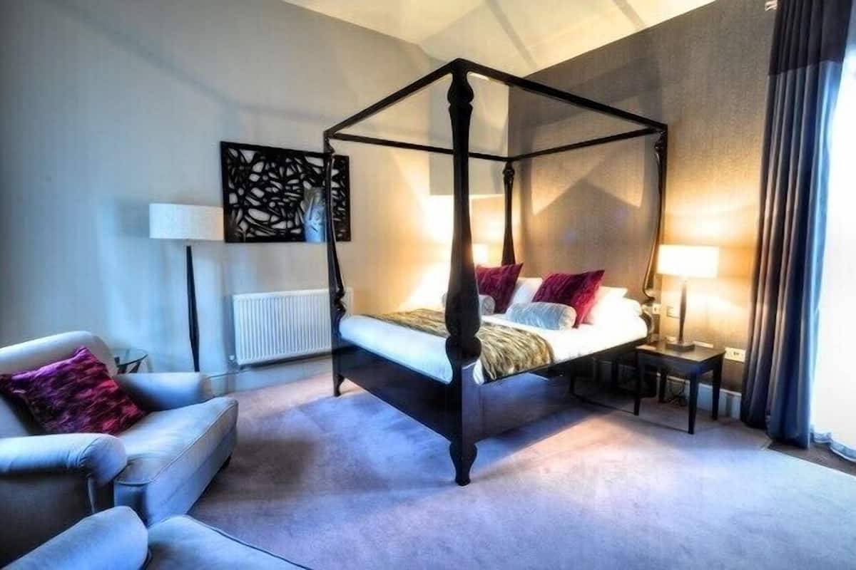 Uma cama com dossel, com edredom branco e vermelho, um sofá cinza com almofada roxa e uma obra de arte abstrata na parede, em preto.