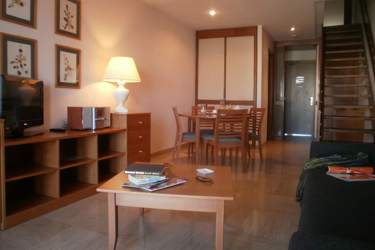 Ein Wohnzimmer mit Holztisch, Stühlen, Fernseher, Lampe und gerahmten Bildern an der Wand.