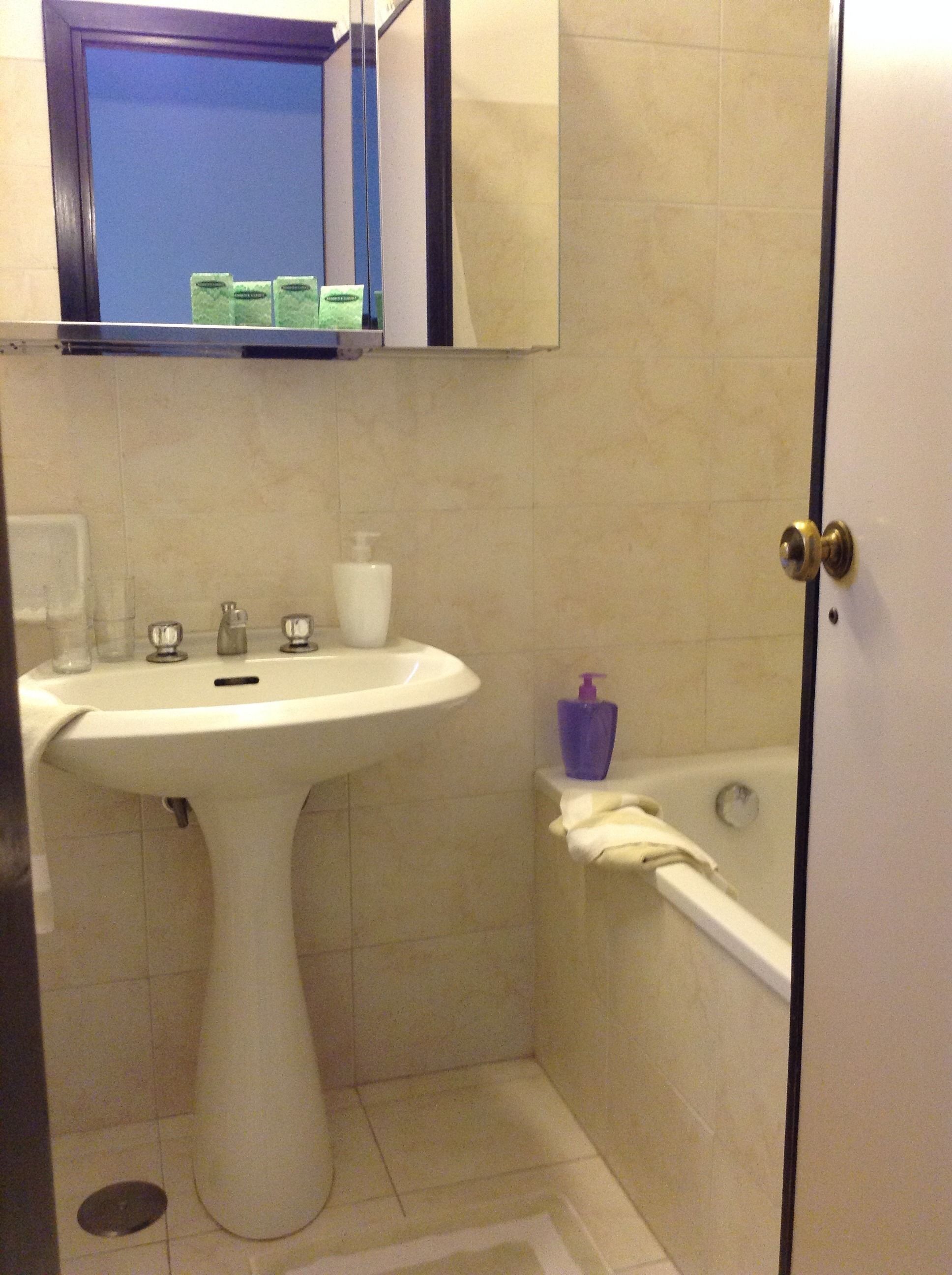 A bathroom with a pedestal sink, a mirror, a bathtub, and a door.