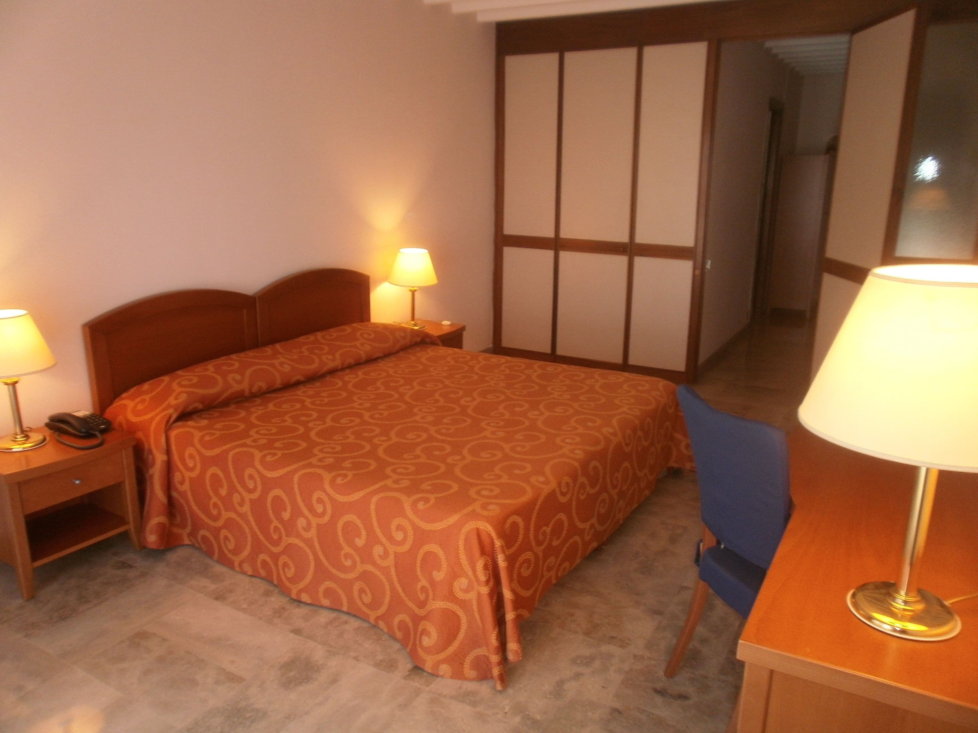 A hotel room with a bed, bedside lamps, a desk with a chair, and a wardrobe.