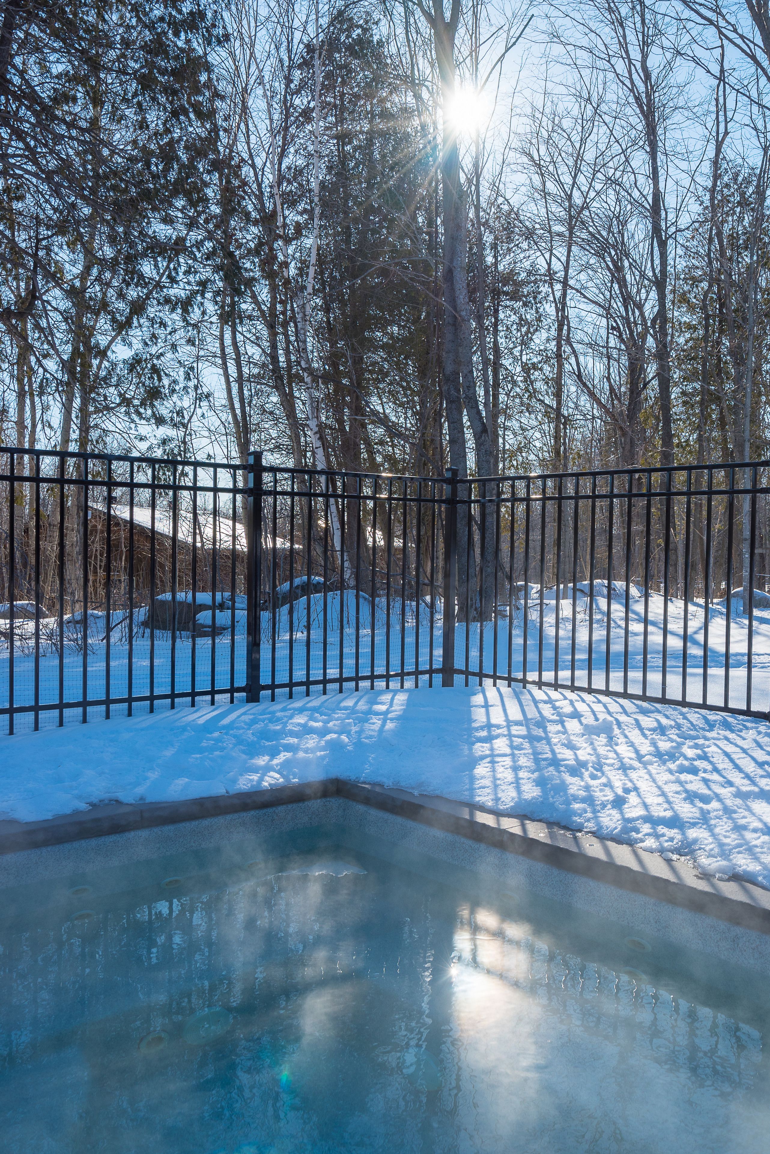 A snow-covered outdoor area with a metal fence, trees, and a partially frozen pool.