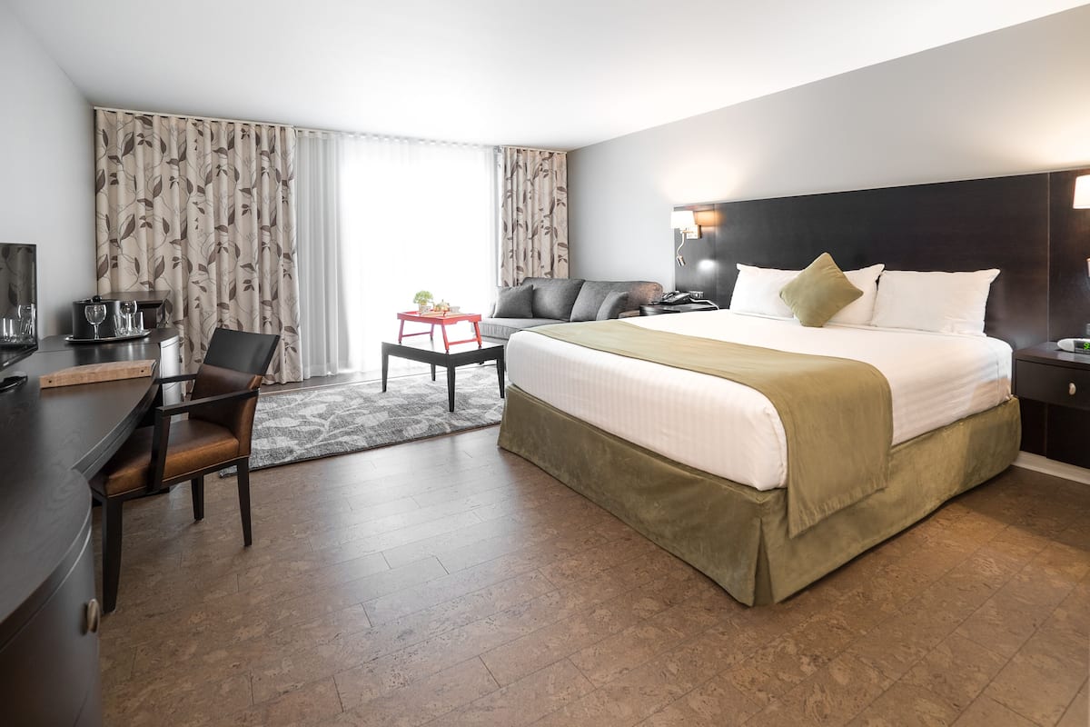 A hotel room with a large bed, a desk with a chair, a small table with a plant, and a window with curtains.