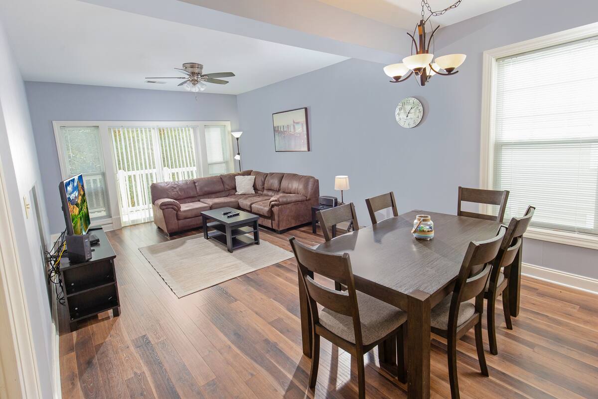 A living room with a sectional sofa, a dining table with chairs, a ceiling fan, and a chandelier.