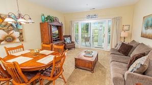 A living room with a wooden dining table, chairs, a sofa, and a coffee table. There is a TV stand with a television and a cabinet with decorative items. A sliding glass door leads to an outdoor area.