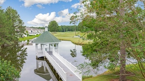 A gazebo with a dock extending into a calm lake, surrounded by lush greenery and a golf course in the background.