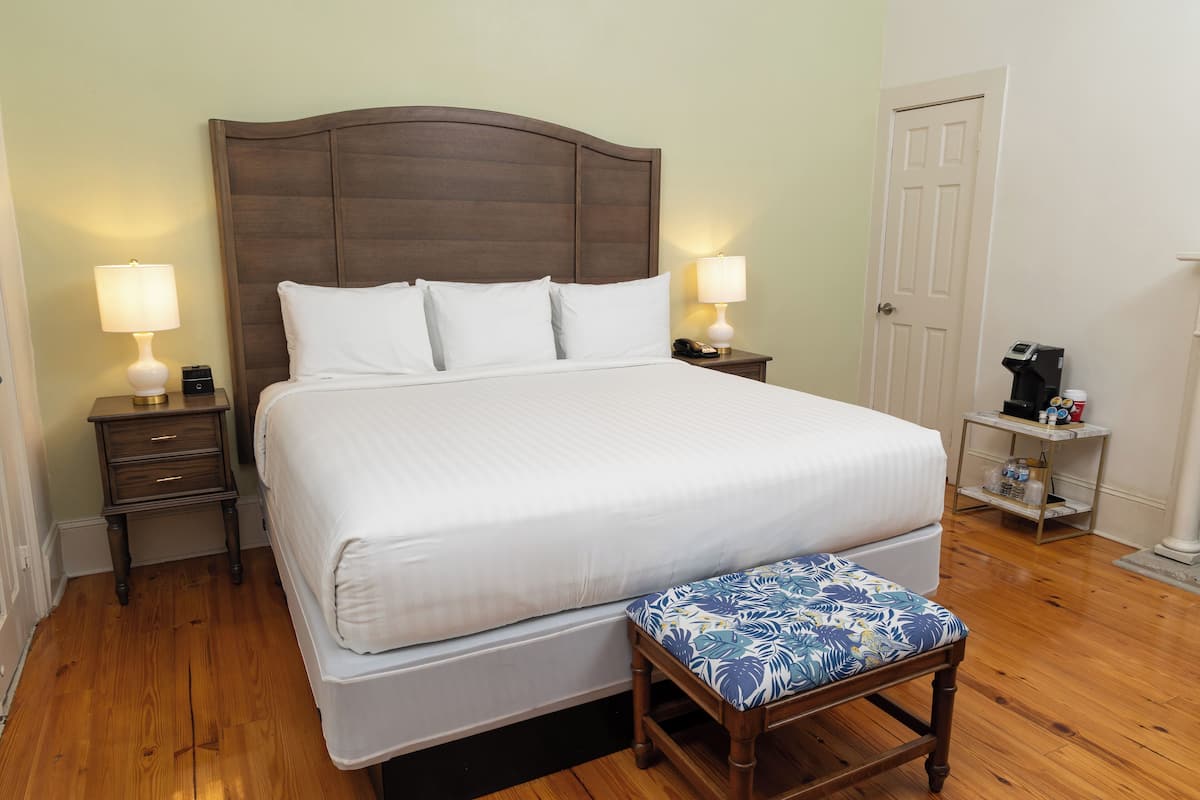 Uma cama bem arrumada com lençóis brancos, duas mesas de cabeceira com abajures, um banquinho de madeira com almofada floral e uma máquina de café sobre uma mesinha.
