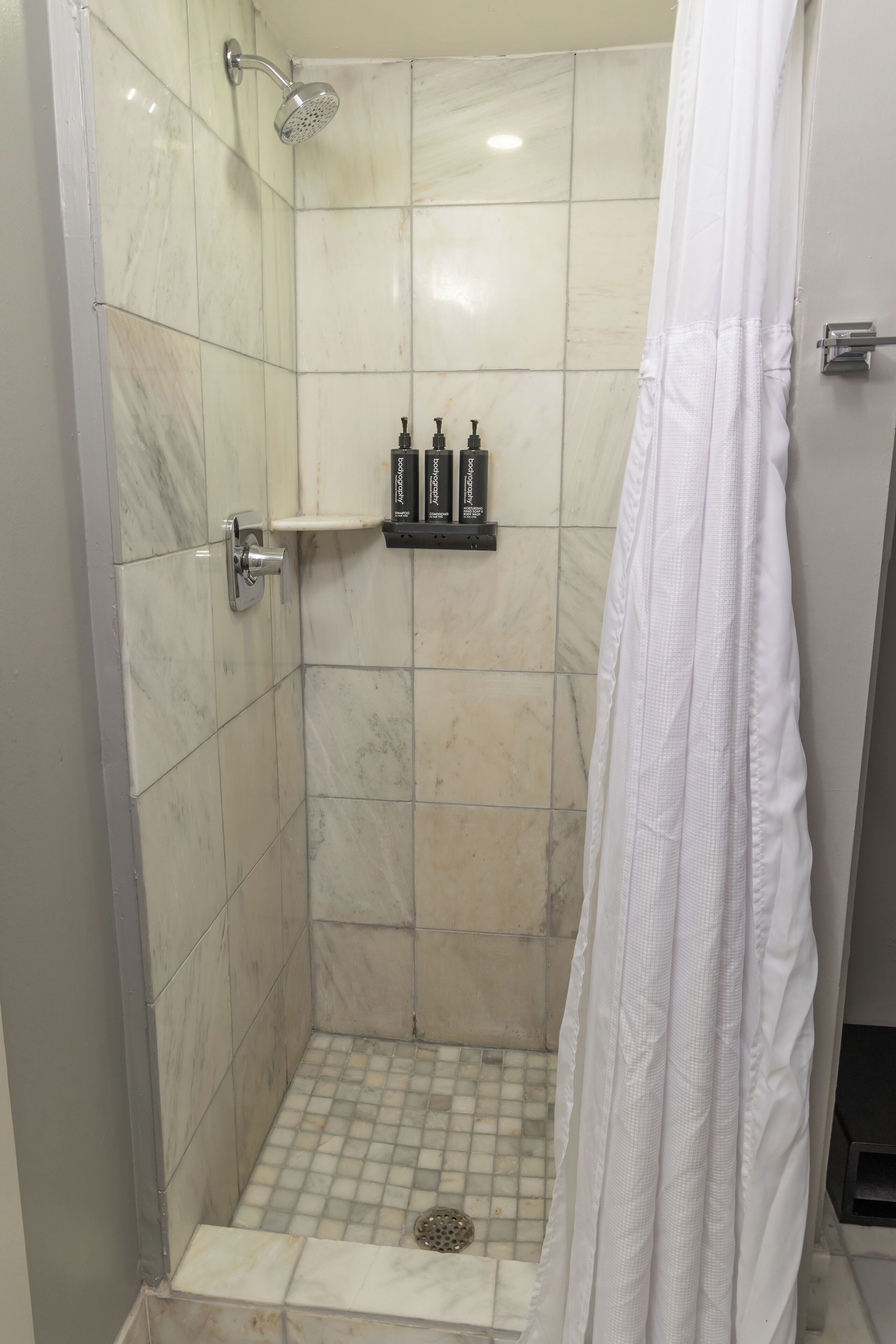 A shower area with a tiled floor, a showerhead, a shelf with toiletries, and a white shower curtain.