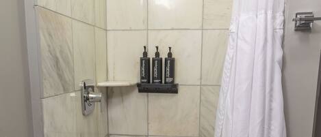 A shower area with a tiled floor, a showerhead, a shelf with toiletries, and a white shower curtain.