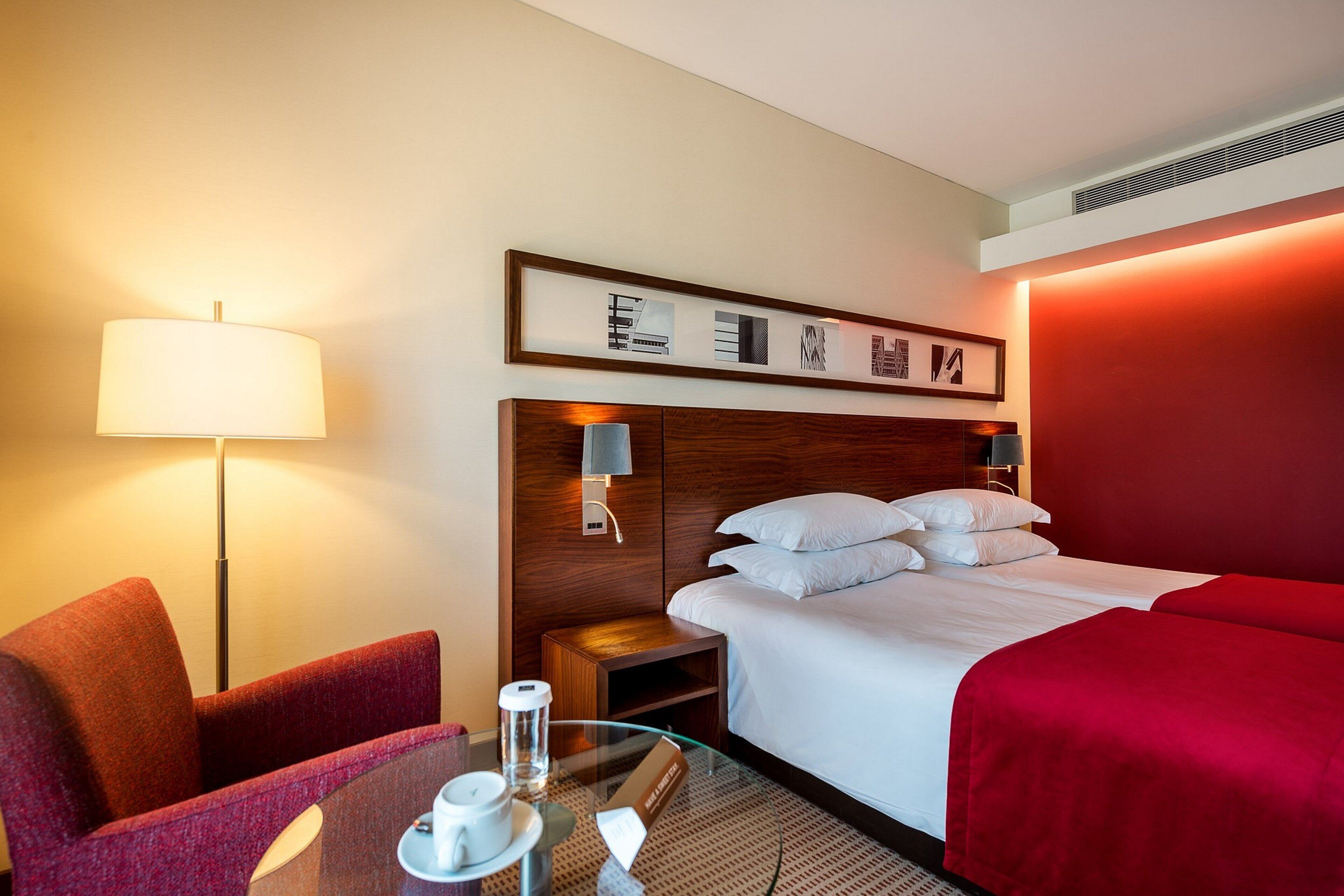 Habitación de hotel con cama, escritorio con lámpara, silla y mesita de noche con un vaso de agua.