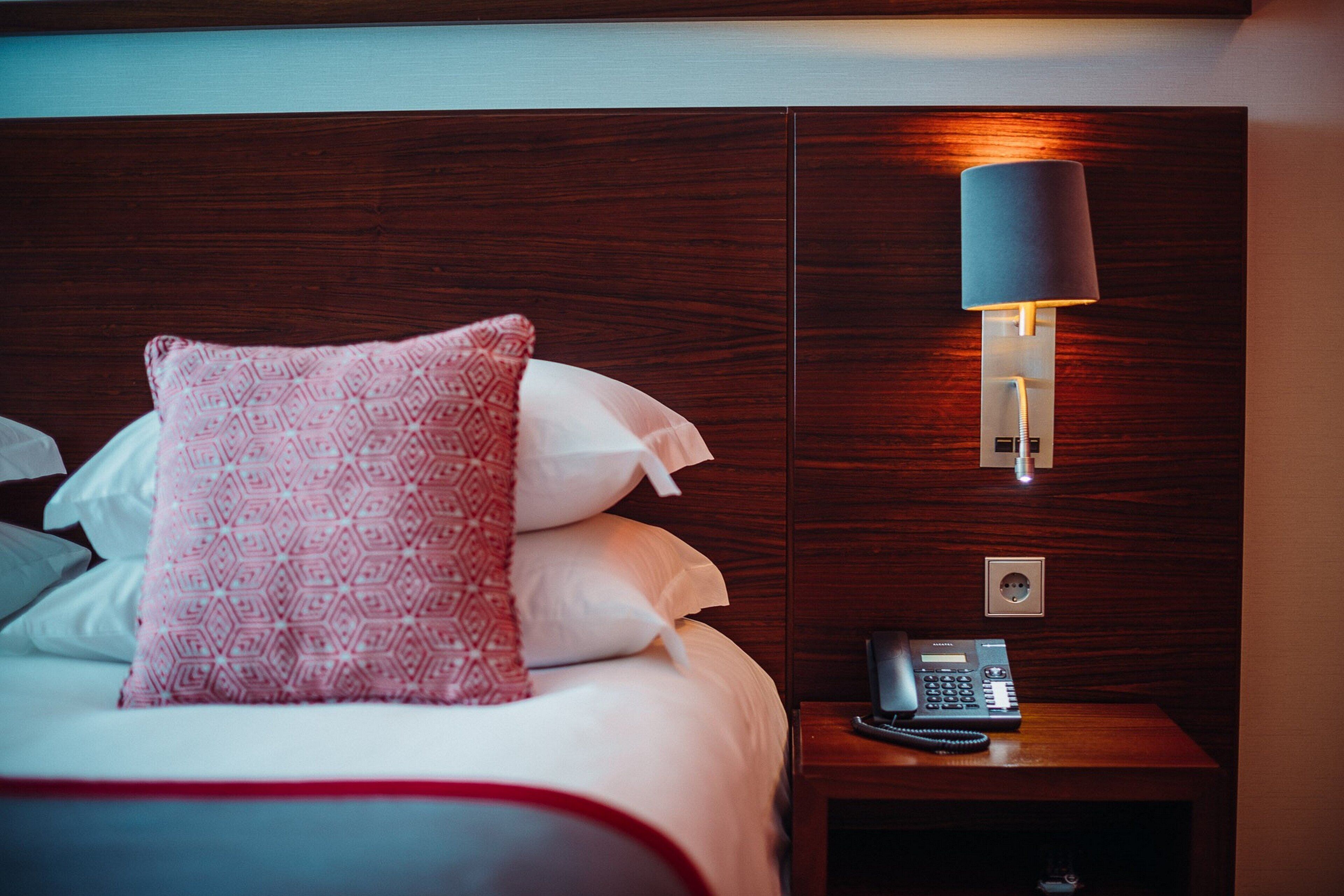 A hotel room with a neatly made bed, a bedside telephone, and a wall-mounted lamp.