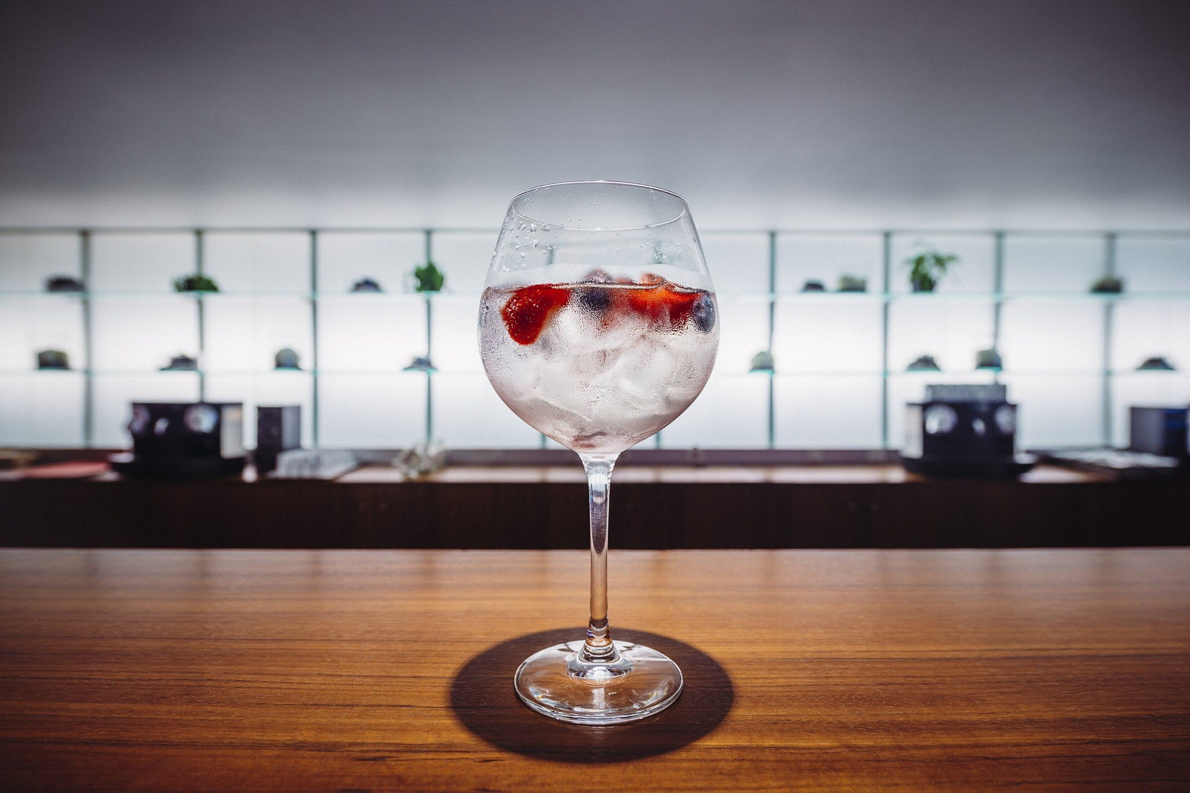 A glass of iced beverage with fruit on a wooden table in a room with a white wall and shelves in the background.
