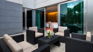 Et udendørs opholdsområde med fletmøbler og et bord, en glasdør der fører til et indendørs rum, og udsigt til grønne områder gennem glasset.