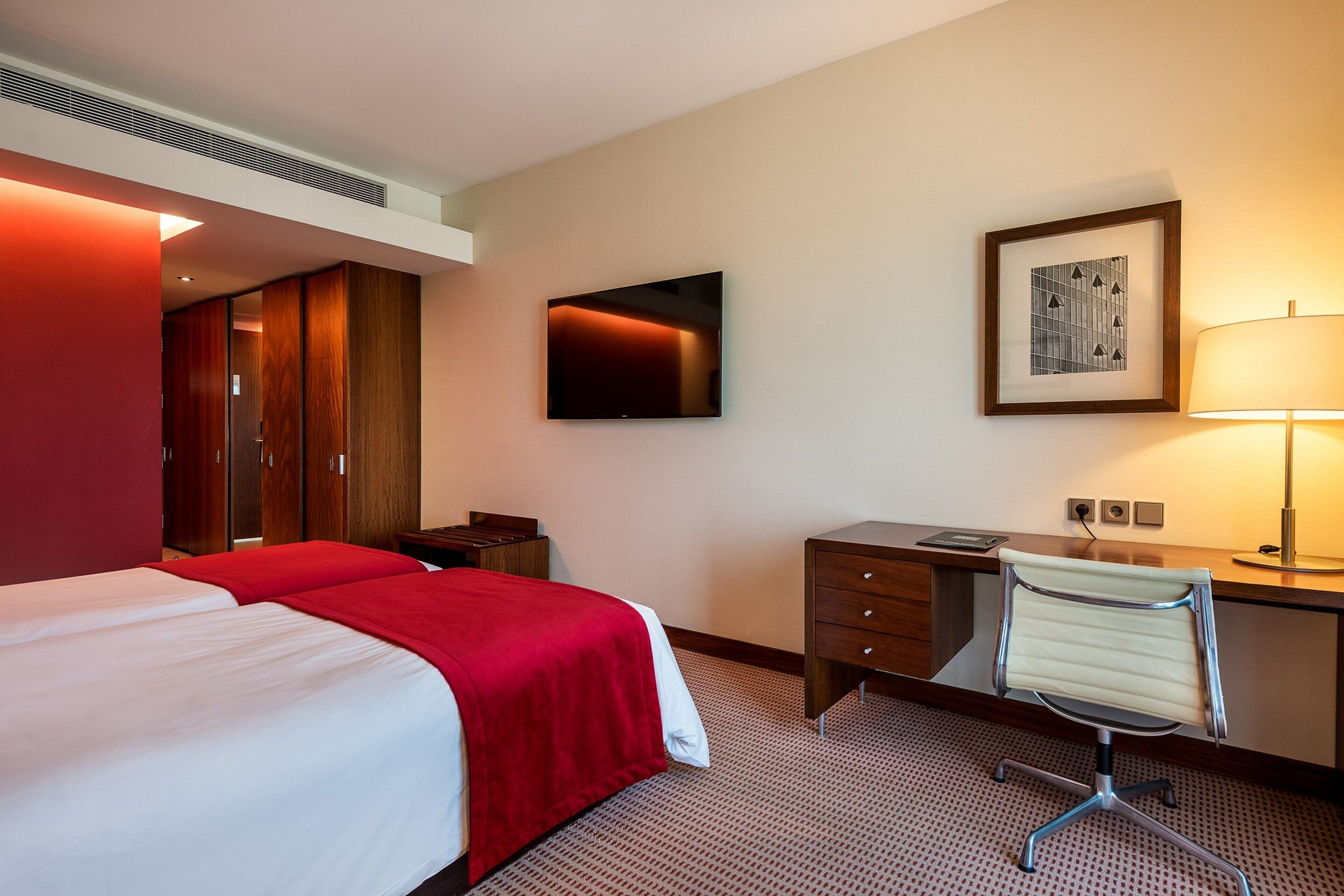 Habitación de hotel con cama, escritorio, silla, televisor, lámpara y un cuadro en la pared.