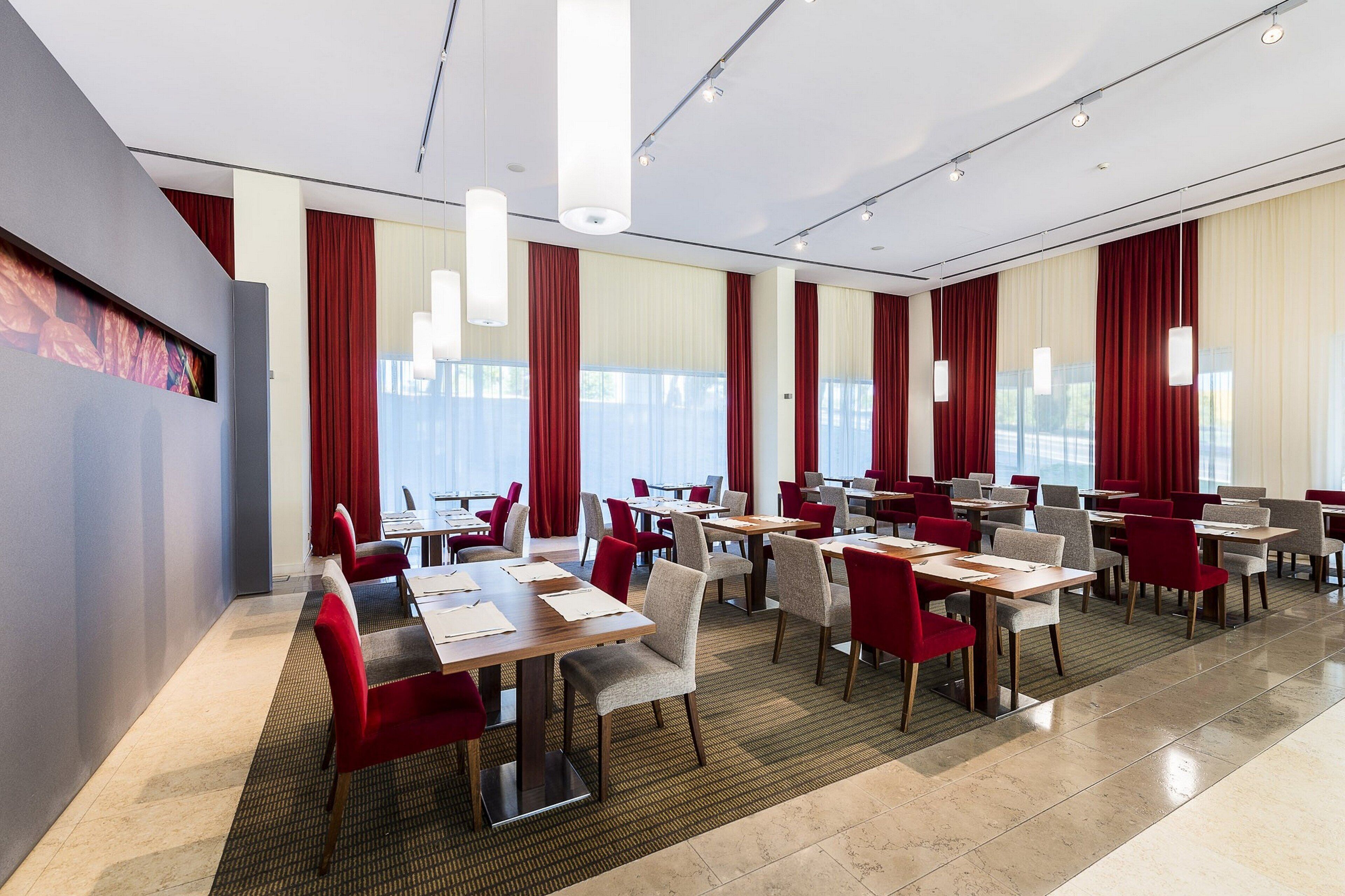 Ein moderner Hotelkonferenzraum mit mehreren Tischen und Stühlen, großen Fenstern mit roten Vorhängen und einem hellen, sauberen Innenraum.