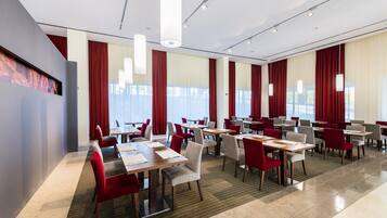 Ein moderner Hotelkonferenzraum mit mehreren Tischen und Stühlen, großen Fenstern mit roten Vorhängen und einem hellen, sauberen Innenraum.