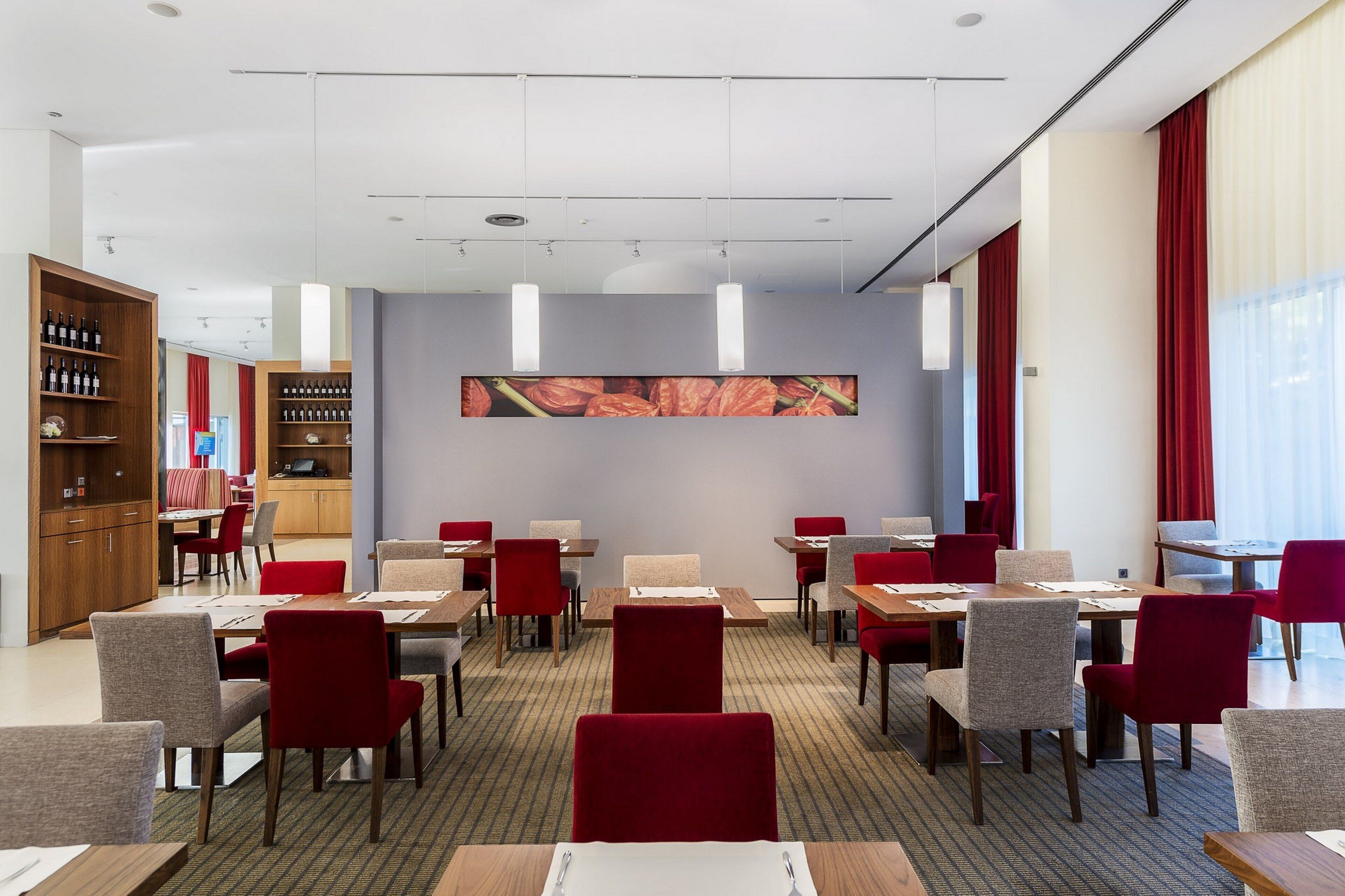 A hotel dining area with multiple tables and chairs, a kitchen area, and a large wall-mounted artwork.