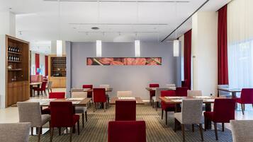 A hotel dining area with multiple tables and chairs, a kitchen area, and a large wall-mounted artwork.