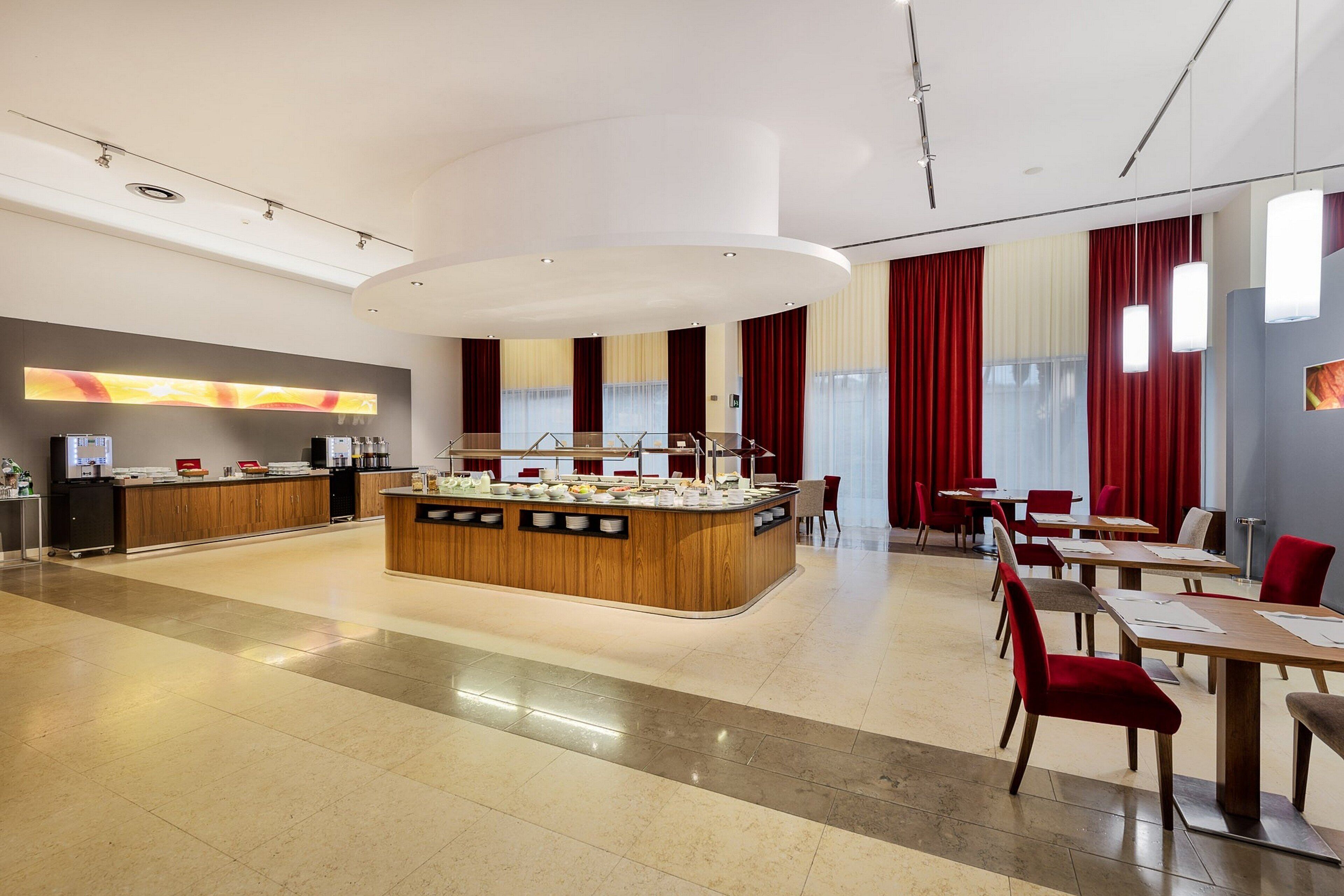 A modern hotel lobby with a central breakfast bar, seating area, and a large window with red curtains.