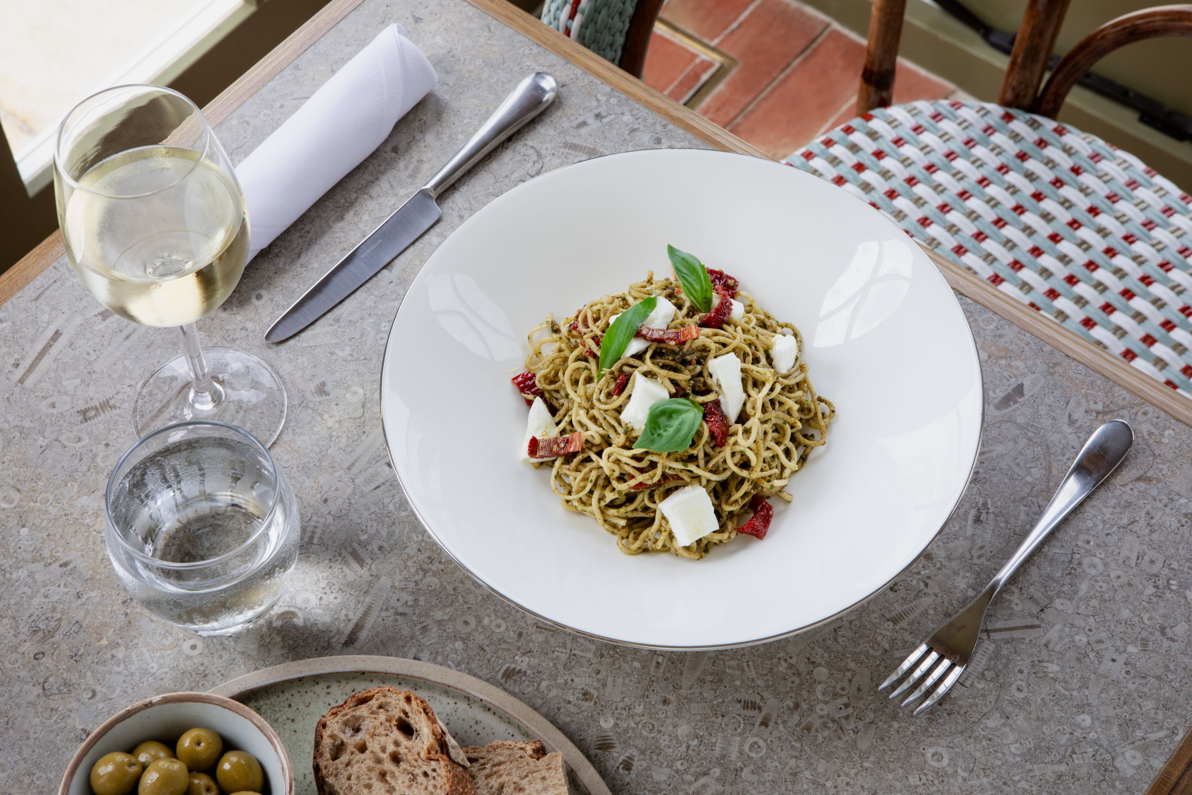 A table with a plate of pasta, a glass of white wine, a bowl of olives, and a loaf of bread.