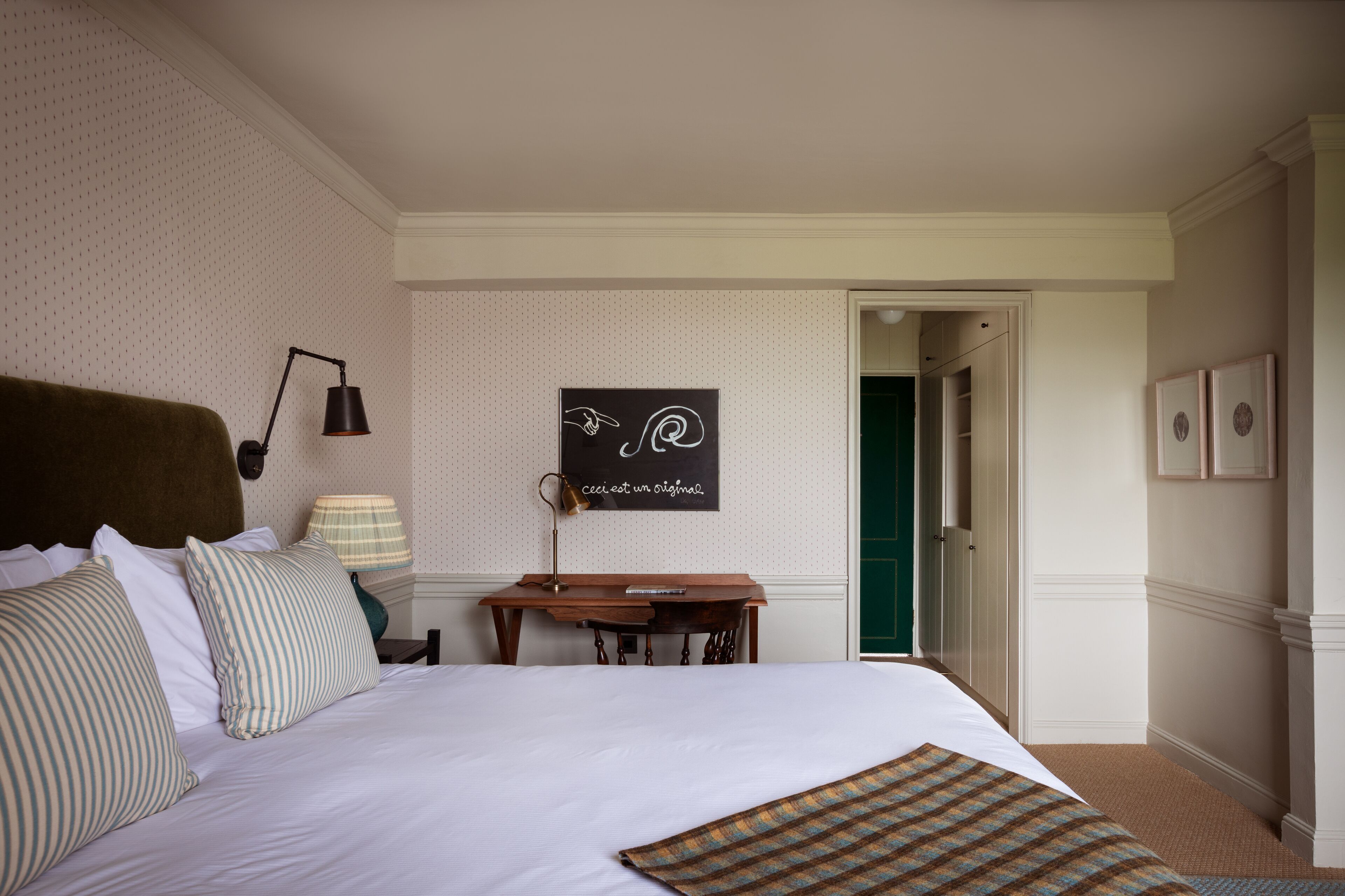 A neatly arranged hotel room with a large bed, a desk with a lamp, a framed quote on the wall, and a door leading to another room.