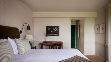 A neatly arranged hotel room with a large bed, a desk with a lamp, a framed quote on the wall, and a door leading to another room.