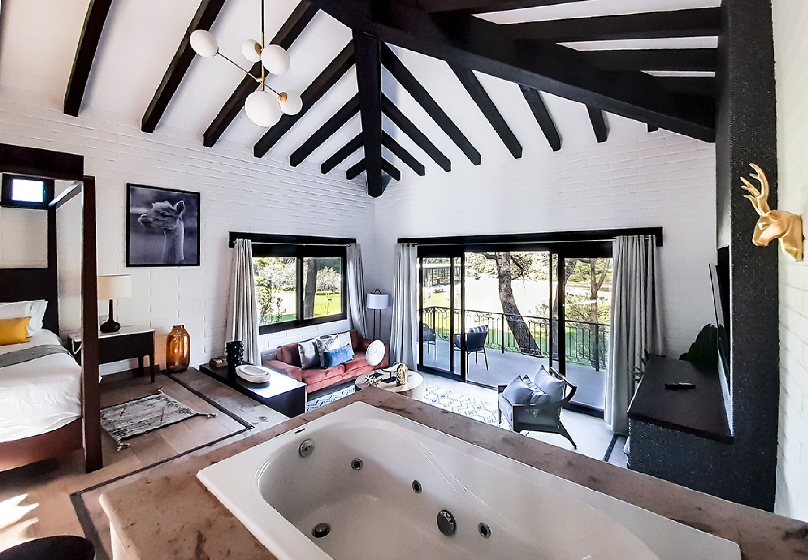 A modern hotel room with a large bathtub, a sofa, a dining table, and a balcony with a view of the outdoors.