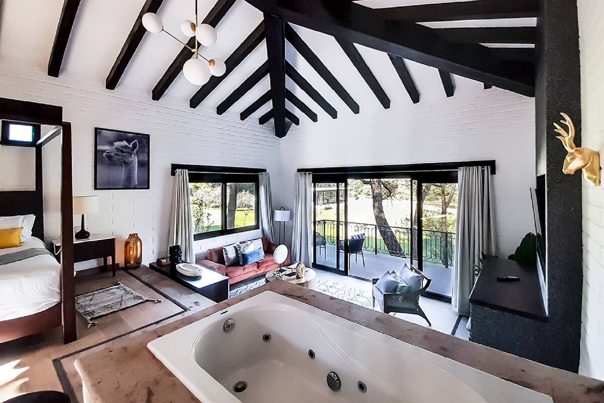 A modern hotel room with a large bathtub, a sofa, a dining table, and a balcony with a view of the outdoors.