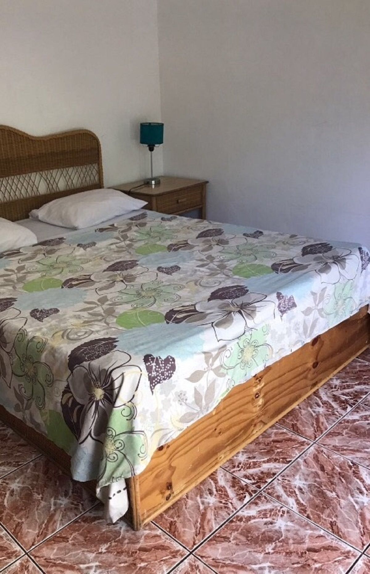 Een bed met een bloemenprint beddegoed, een houten hoofdbord, een nachtkastje met een lamp en een betegelde vloer.