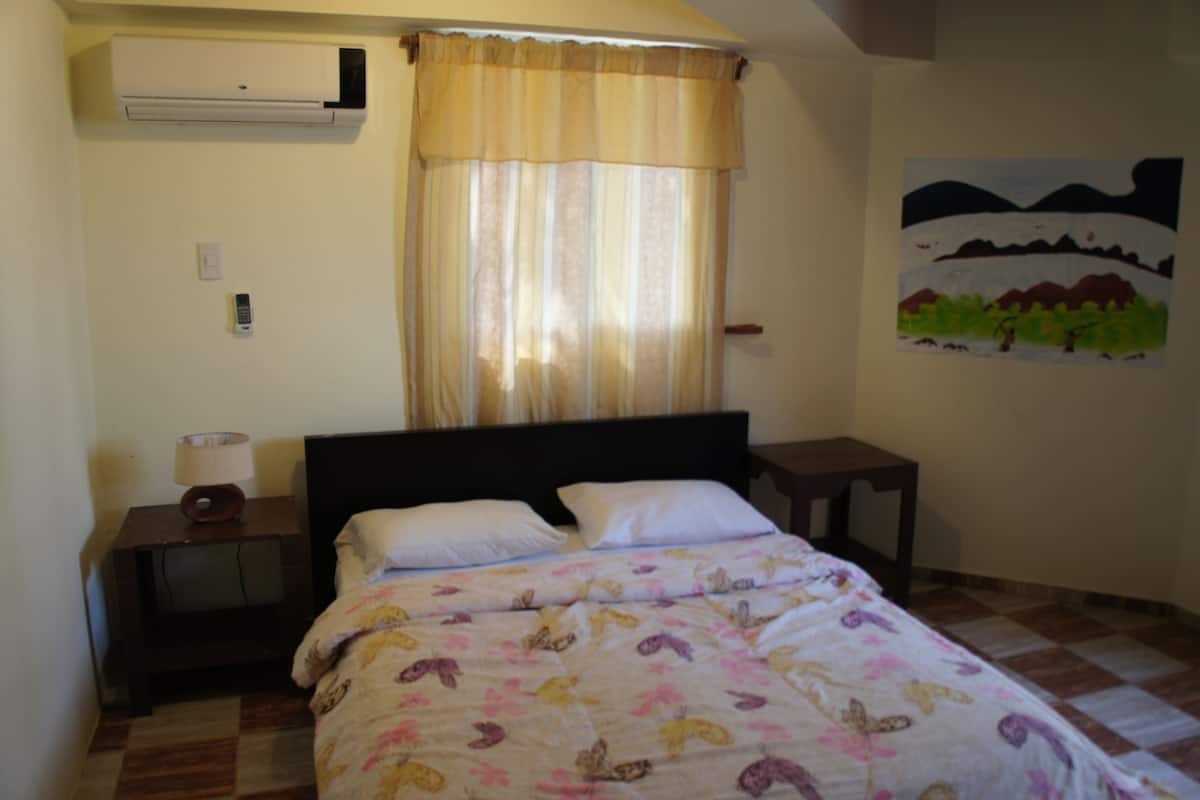 A bedroom with a bed, a nightstand with a lamp, a small table, a wall-mounted air conditioner, and a painting on the wall.
