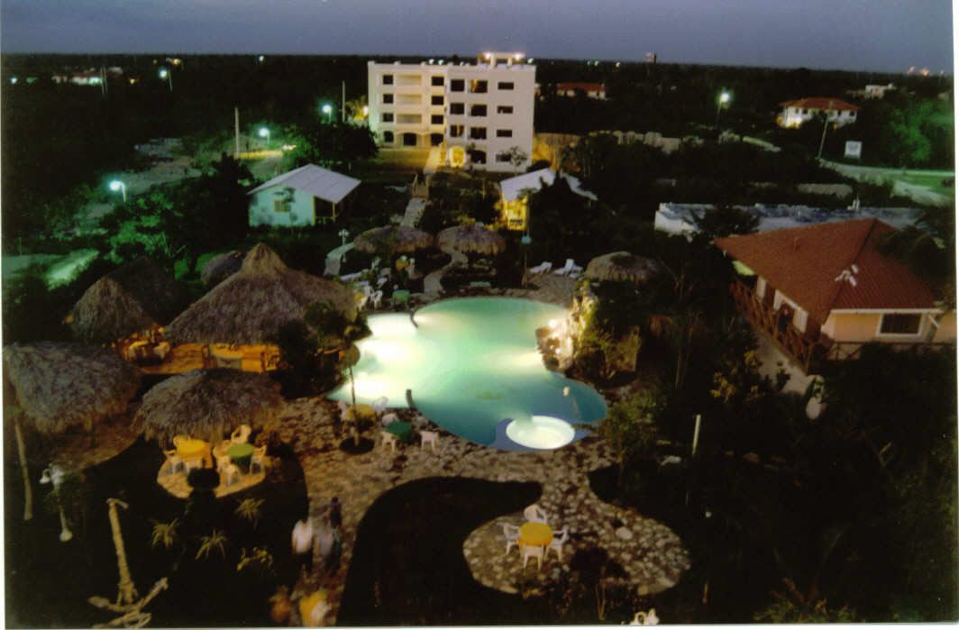 Un resort con una piscina central, estructuras de techo de paja y un edificio grande al fondo.